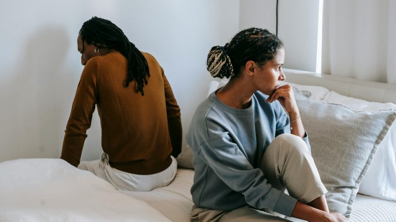 Unhappy couple sitting on a bed facing away from each other, looking upset, in a softly lit bedroom.