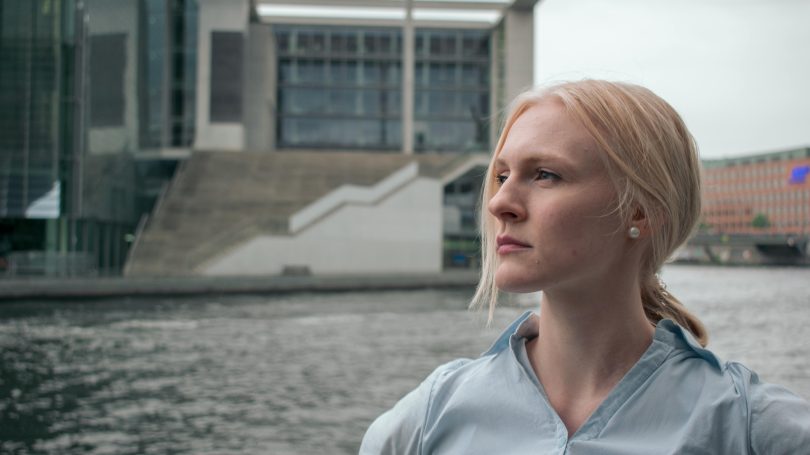 A determined woman with blonde hair, wearing a light blue shirt, gazes into the distance by a river, with modern architecture in the background