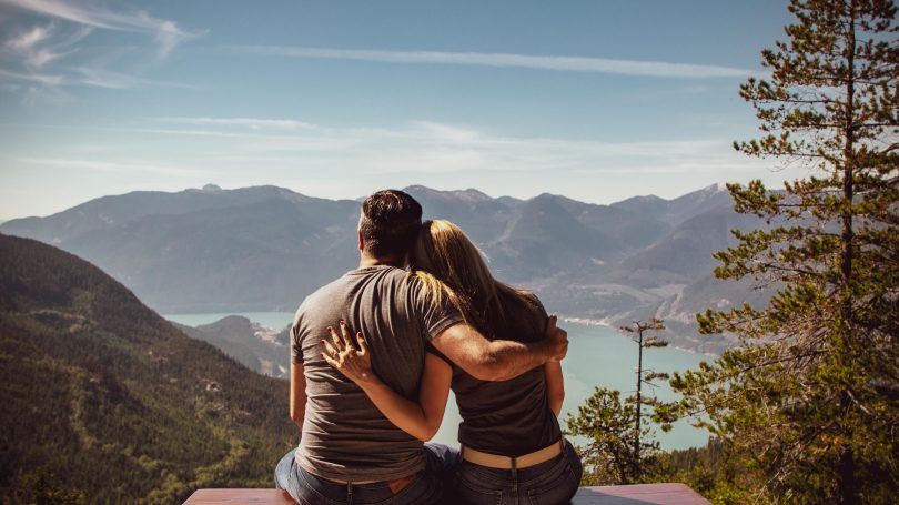 A couple sitting on a wooden bench, embracing while looking at a scenic mountain and lake view.