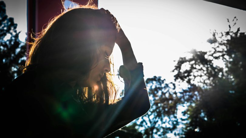 A woman standing outdoors with her hand on her head, looking distressed as sunlight shines behind her, creating a dramatic effect.