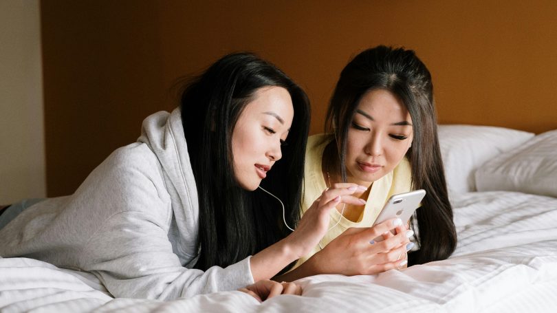 Two young women lying on a bed, sharing earphones and looking at a smartphone together.