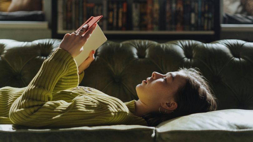 Young woman lying on a green couch, reading a book in a cozy living room setting.