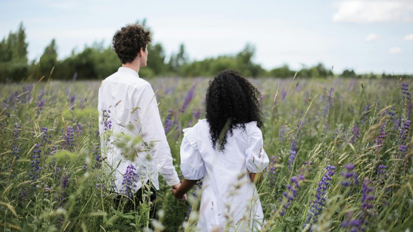 A couple holding hands while walking through a field of tall grass and purple wildflowers.