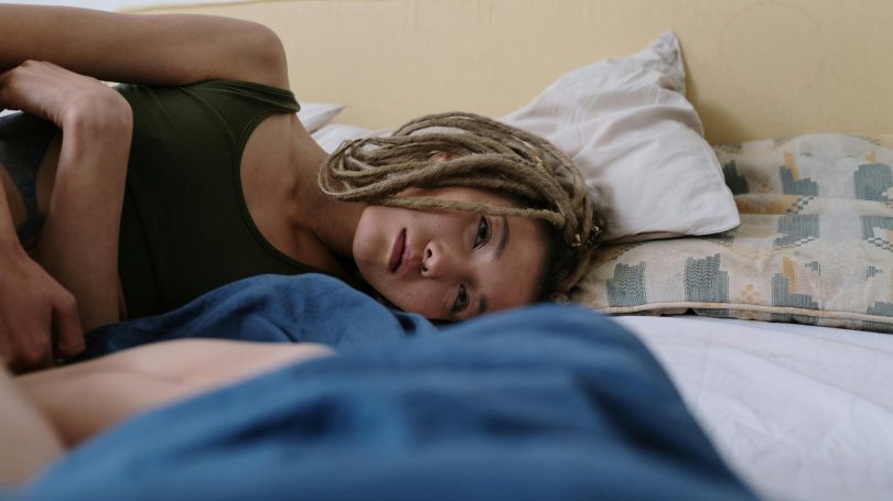A woman with dreadlocks lying on her side in bed, gazing into the distance with a pensive expression. Soft natural lighting enhances the intimate and reflective mood.