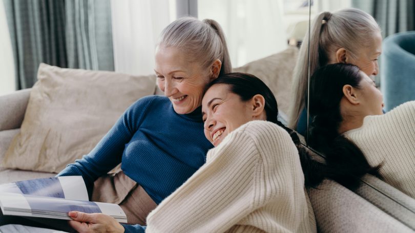 An older woman with her arm around a younger woman are sharing a laugh while reading a magazine together on the sofa.