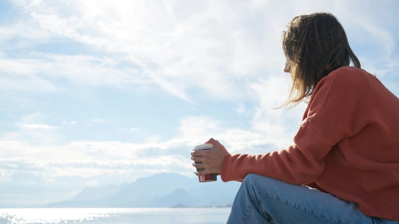 Young woman sitting by the sea, holding a coffee cup, and reflecting on health and lifestyle choices
