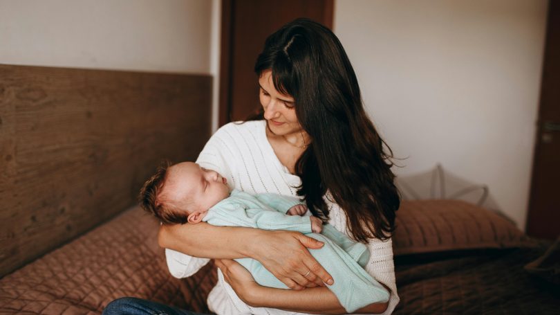 A new mother sitting by the window, looking thoughtful while holding her baby, representing postpartum emotions and the baby blues.