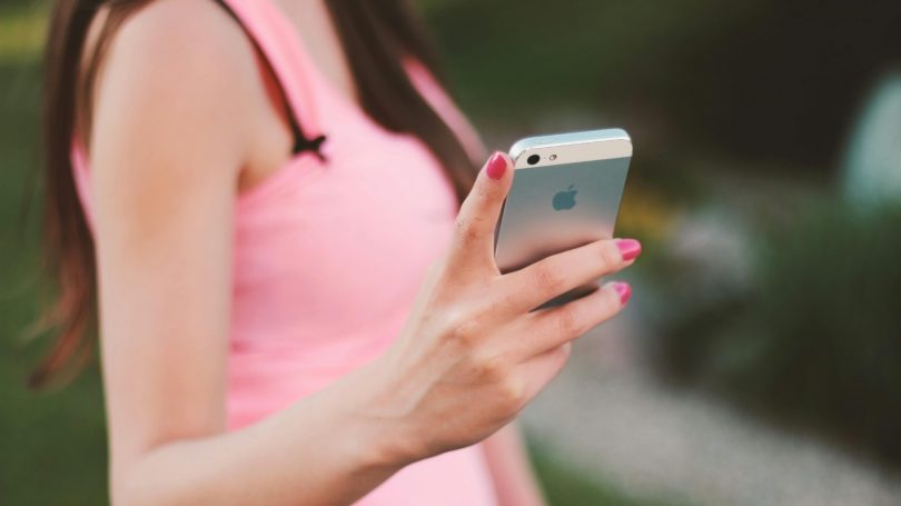 Woman in a pink tank top holding a silver iPhone outdoors with a blurred background.