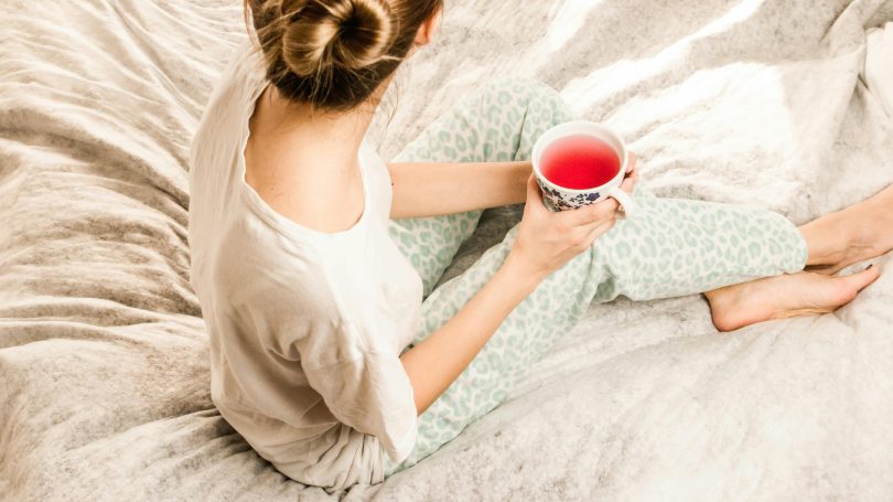 Woman sitting on a bed in cozy pajamas, holding a cup of red herbal tea, with her hair in a bun, enjoying a quiet and relaxed moment.