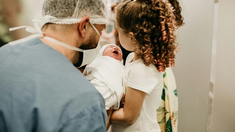 Doctor in surgical attire holding a newborn baby while a young girl with curly hair meets the baby for the first time in a hospital setting.