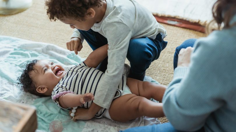 Young child playfully tickling a laughing baby on a soft blanket while a parent watches nearby.