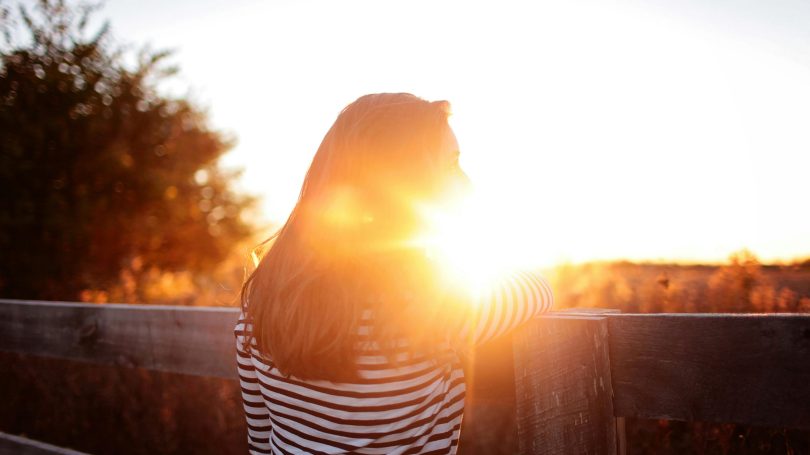 A woman in a striped shirt leaning on a wooden fence, gazing into the sunset with a warm glow surrounding her.