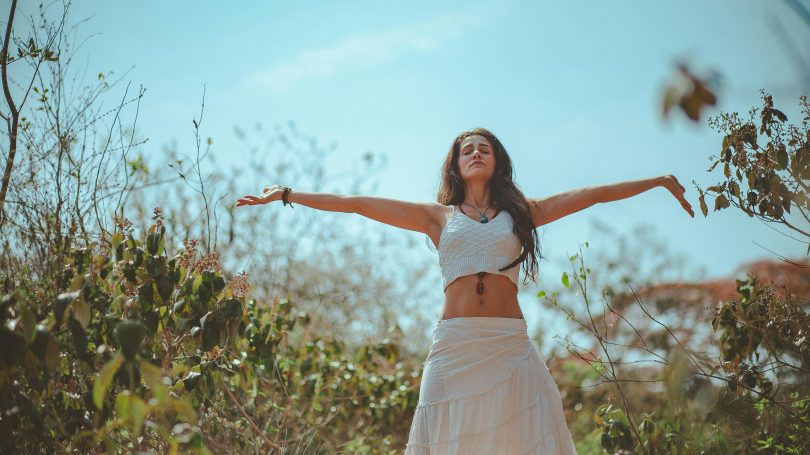 A woman standing outdoors with arms outstretched, eyes closed, and an expression of calm. She is wearing a white crop top and a flowing white skirt, surrounded by green foliage under a bright blue sky