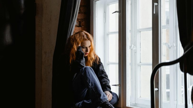 Young woman with red hair sitting by a window, looking pensive in a dimly lit room.