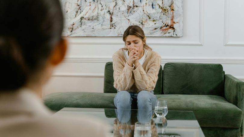 A distressed woman sitting on a green couch, holding her hands near her face, appearing emotional while speaking to a therapist in a professional setting.