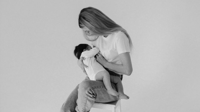 A mother sitting on a stool, lovingly breastfeeding her baby while looking down at them in a peaceful, minimalistic setting.