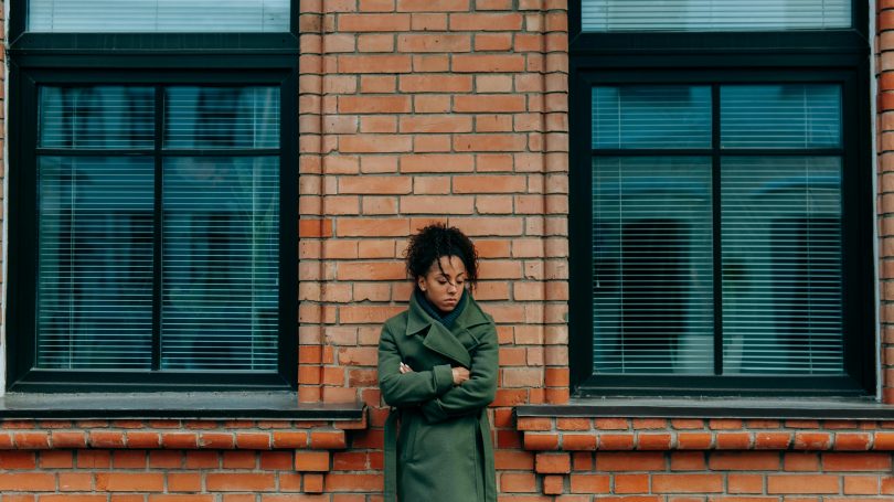 A woman in a green coat standing against a brick wall with her arms crossed, looking down with a contemplative expression, symbolizing sadness or deep thought.