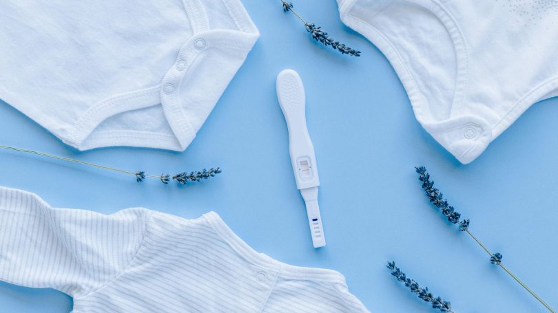 A positive pregnancy test placed on a blue background, surrounded by white baby clothes and lavender sprigs.