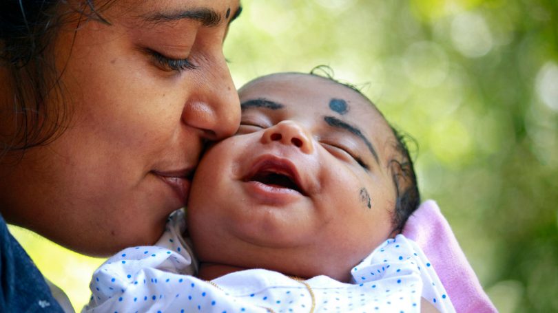 A mother lovingly kisses her smiling baby, who is dressed in a white polka-dotted outfit.