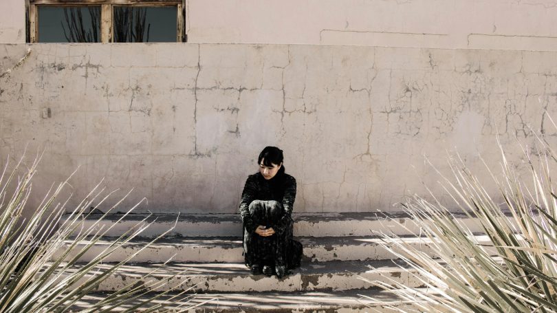 A woman dressed in black sits alone on worn concrete steps, looking down in contemplation against a cracked wall, symbolizing isolation and emotional distress.