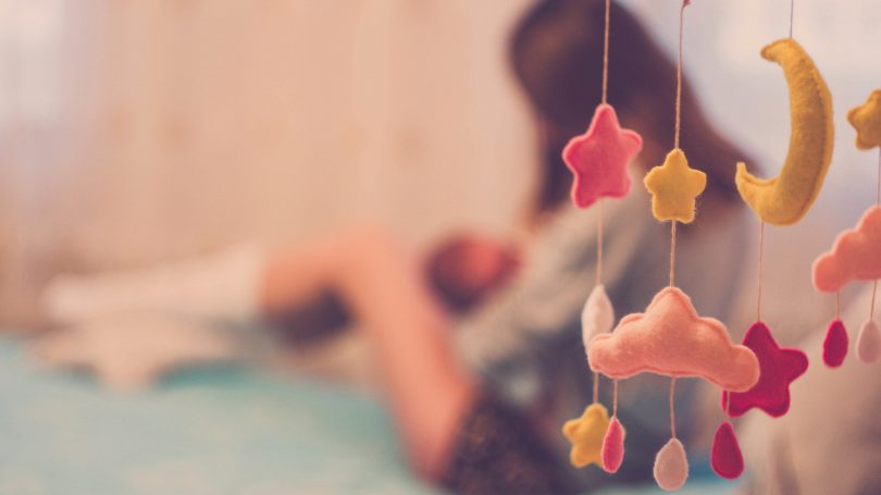 A soft-focus image of a mother holding her baby in the background with a hanging baby mobile in the foreground, symbolizing the emotional journey of postpartum motherhood.