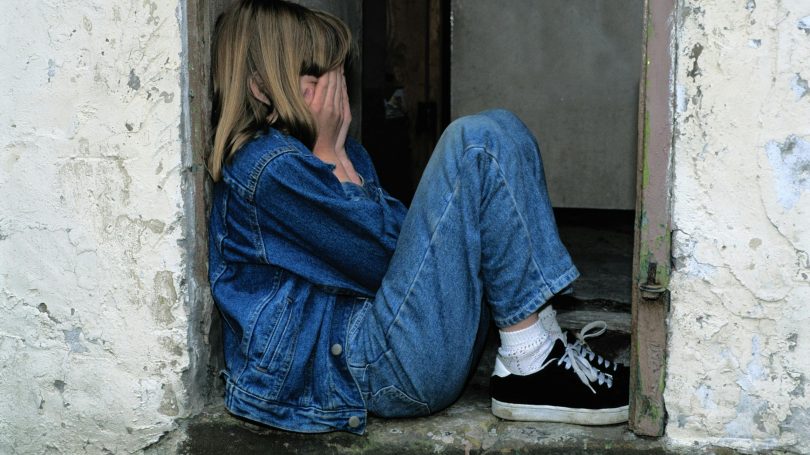 Sad young girl in a denim jacket sitting on a worn doorstep, covering her face with her hands.