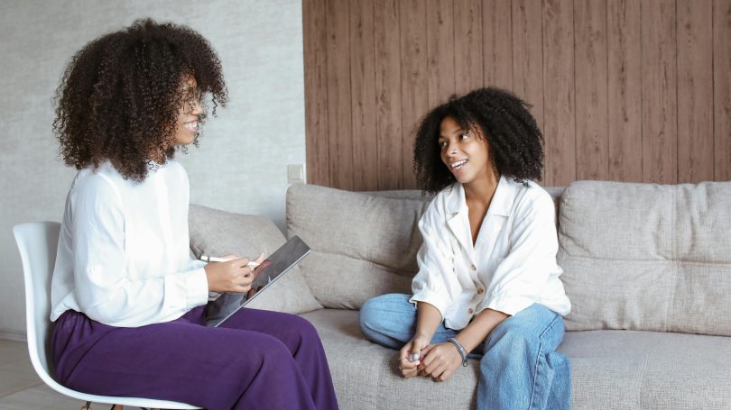 Smiling therapist holding a tablet while talking with a young woman sitting on a couch during a counseling session.