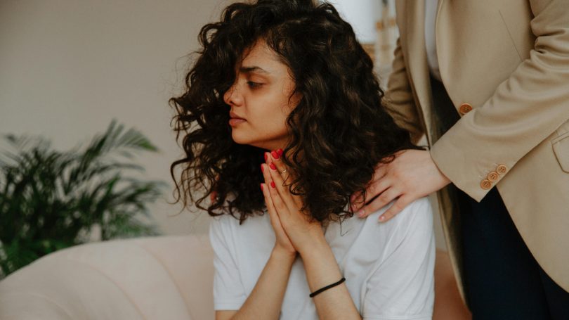 A distressed young woman with curly hair sitting on a couch, clasping her hands together while someone offers comfort by placing hands on her shoulders.