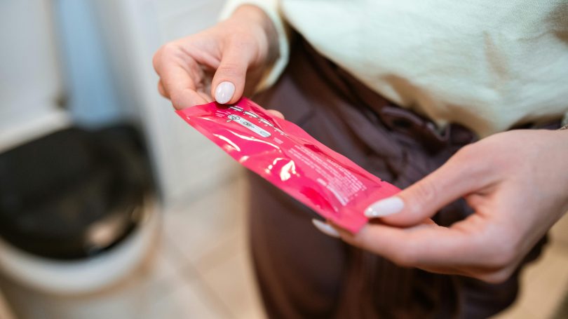 A woman holding a pink pregnancy test package in a bathroom, preparing to take a test.