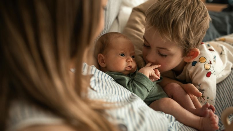 A tender moment between siblings as an older brother gently holds and kisses the tiny hand of his newborn sibling while their mother watches from the foreground.