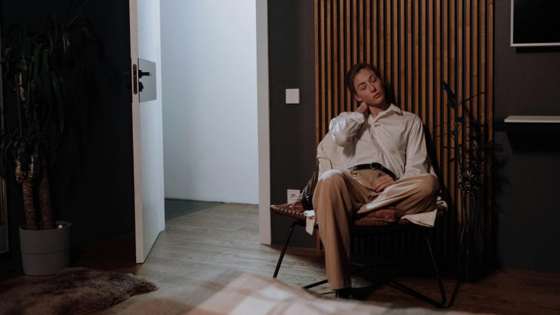 Woman sitting in a dimly lit room, leaning back in a chair with his eyes closed, appearing tired or deep in thought.