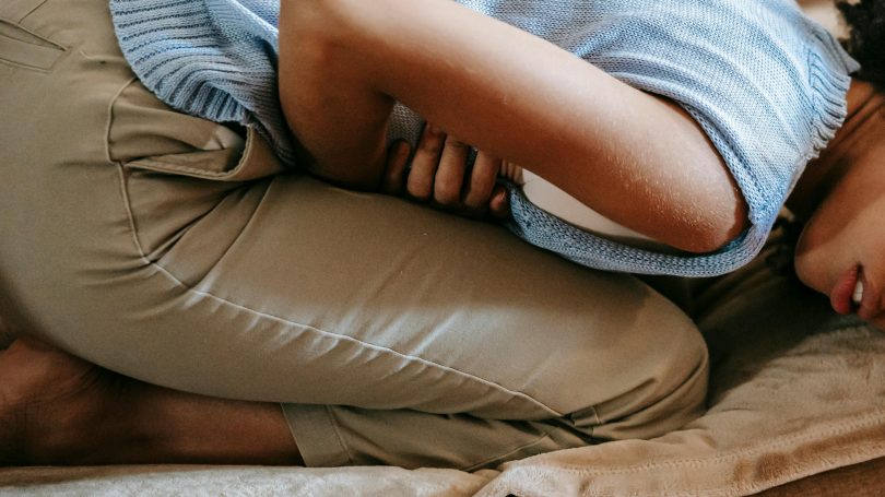 A woman lying on a couch in a fetal position, holding her stomach, expressing pain or discomfort.