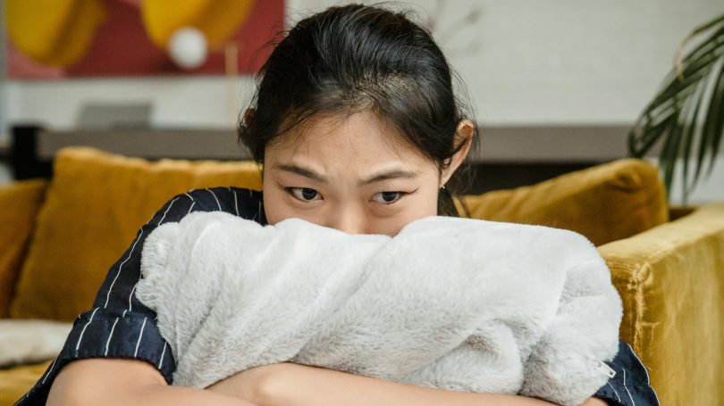 A worried young woman hugging a soft blanket while sitting on a couch, looking anxious and deep in thought.