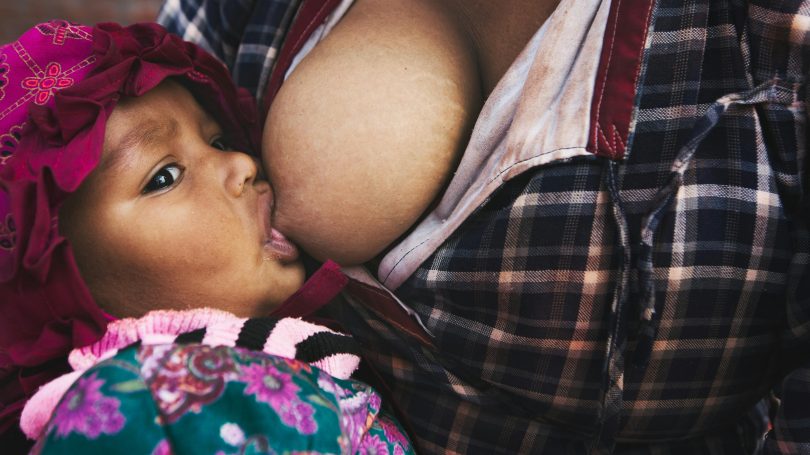 A close-up of a baby breastfeeding, wearing a red bonnet and a floral outfit, while the mother, dressed in a plaid shirt, holds the child close.