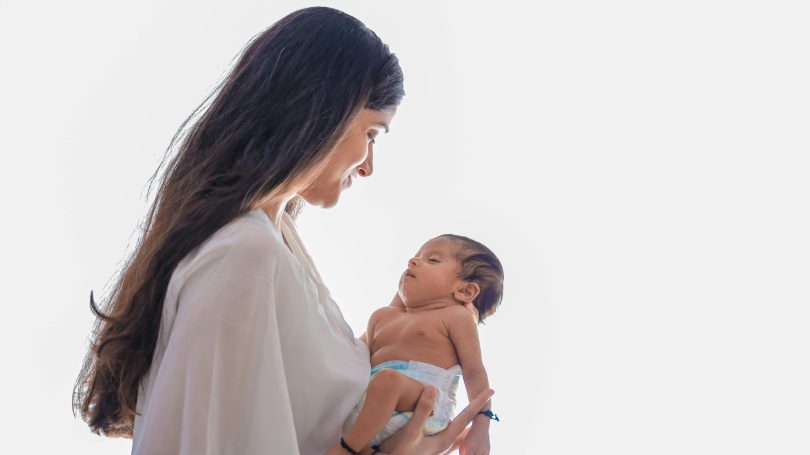 A mother in a white blouse gently cradling her newborn baby while gazing at them with love and care.