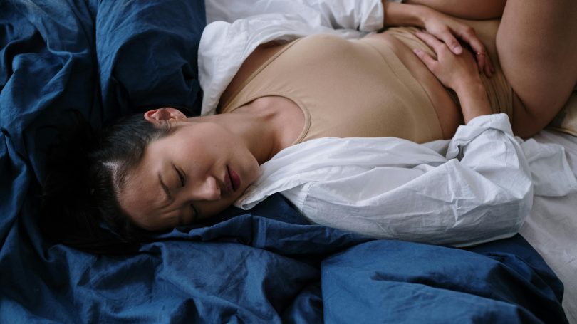 Woman lying in bed, holding her lower abdomen, appearing to be in pain or discomfort.