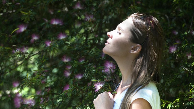 Woman standing outside with eyes closed, facing the sun, surrounded by green leaves and pink flowers.