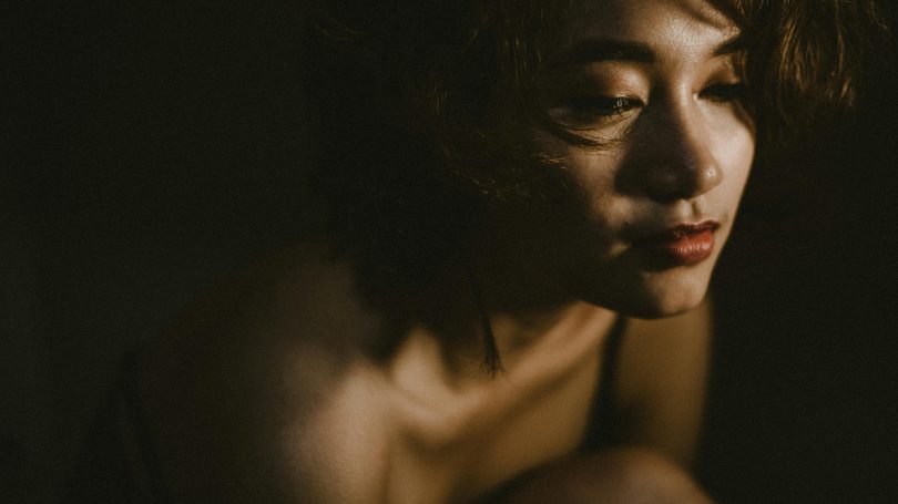 Close-up of a woman in low light, looking down with a somber expression, suggesting deep thought, sadness, or emotional reflection.