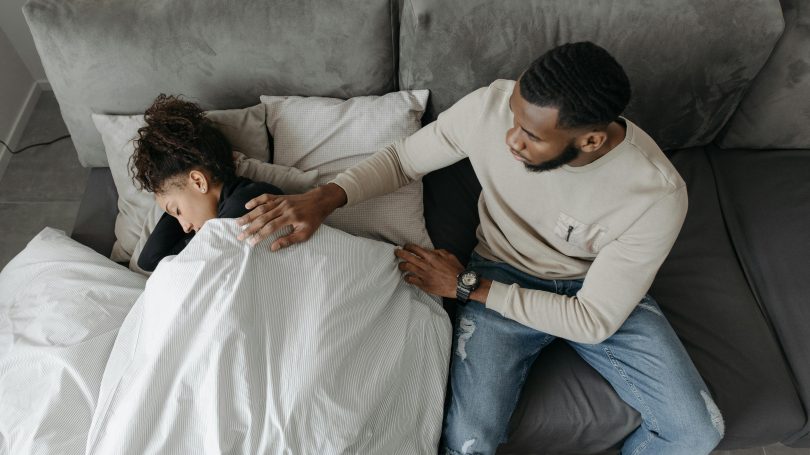 Man sitting beside a woman lying in bed under a blanket, gently placing a hand on her back in a comforting and supportive gesture.