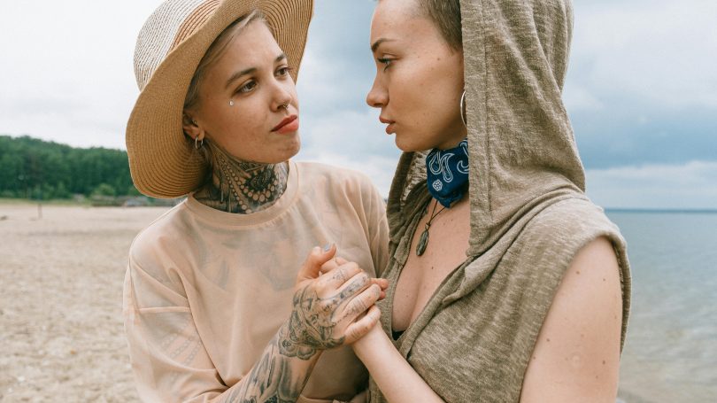 Two women standing close on a beach, holding hands and looking at each other with empathy and support, suggesting a moment of emotional connection.