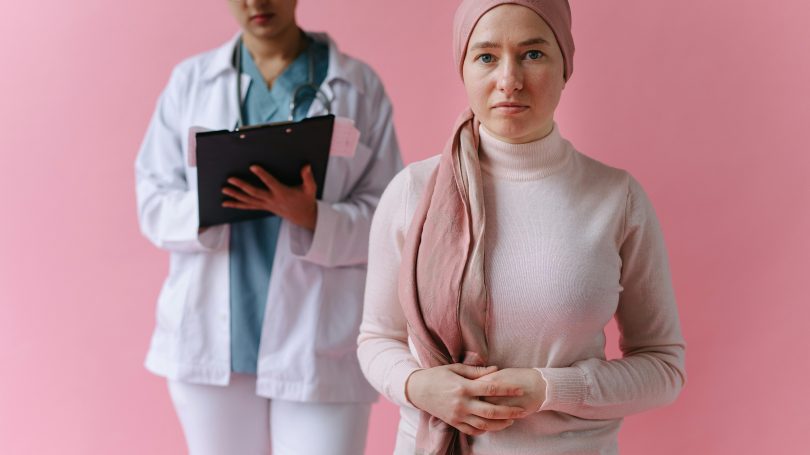 Woman wearing a headscarf and light pink turtleneck standing in front of a healthcare professional holding a clipboard, set against a pink background, suggesting a medical consultation or support setting.